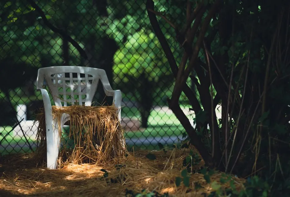 A deteriorating plastic chair surrounded by hay and vegetation near a garden fence.